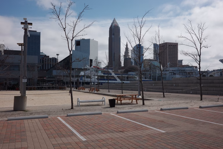 The image shows a view of the Cleveland skyline from North Coast Harbor on a bright, slightly overcast day. In the foreground, a paved parking area with red bricks and white space markers leads to a sandy public square. Several young, leafless trees are scattered across the sand, interspersed with wooden picnic tables and a white bench. The background is dominated by iconic skyscrapers, including the tall, pointed Key Tower, the historic Terminal Tower spire, and the modern 200 Public Square. To the left, a large white boat is docked.