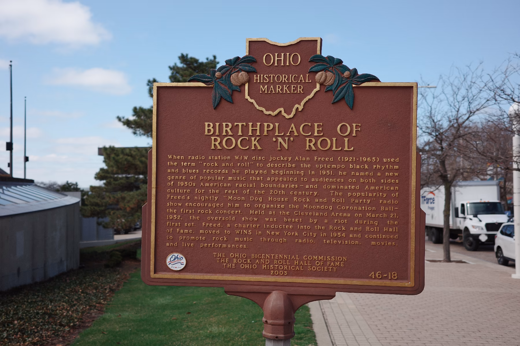 An Ohio Historical Marker stands in the foreground against a clear blue sky. The bronze, rectangular plaque is topped with a decorative "Ohio Historical Marker" emblem featuring a buckeye. Titled "BIRTHPLACE OF ROCK 'N' ROLL," the plaque's gold text explains how Cleveland disc jockey Alan Freed coined the term in 1951 and organized the first rock concert in 1952. The marker is positioned on a grassy area next to a paved walkway. In the soft-focus background, a white truck and modern city buildings are visible under a bright, sunny sky.